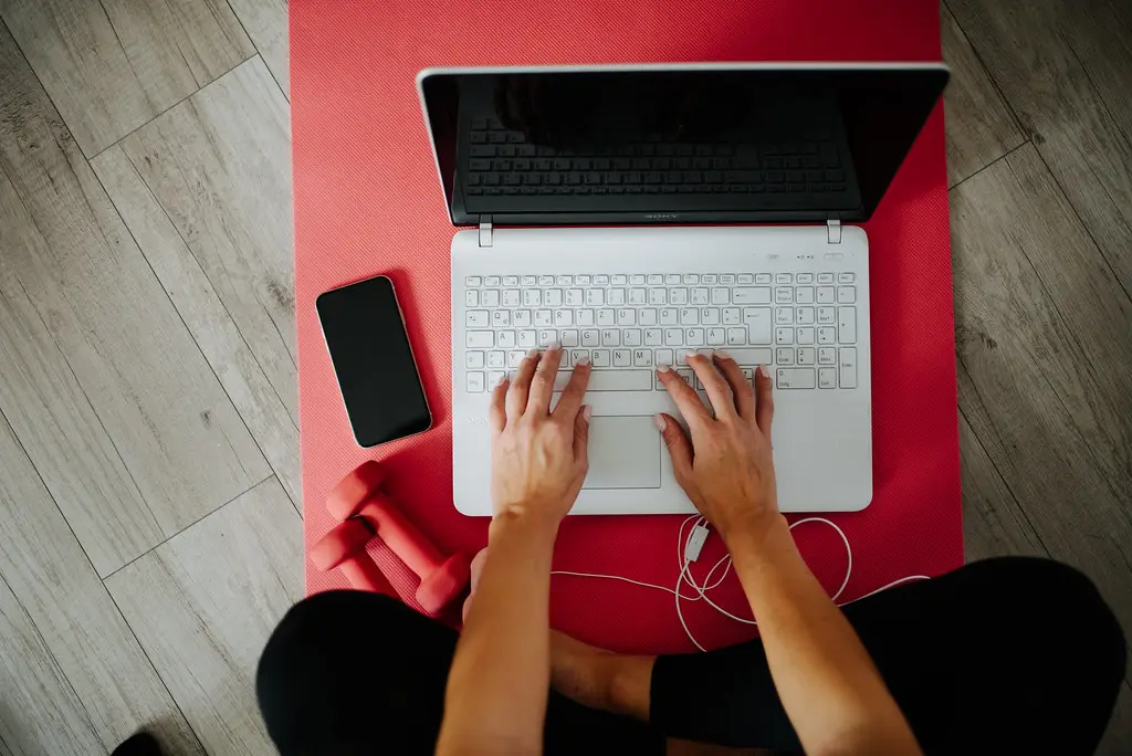 Foto: A person sits cross-legged on a red yoga mat, typing on a laptop with weights and a smartphone nearby
