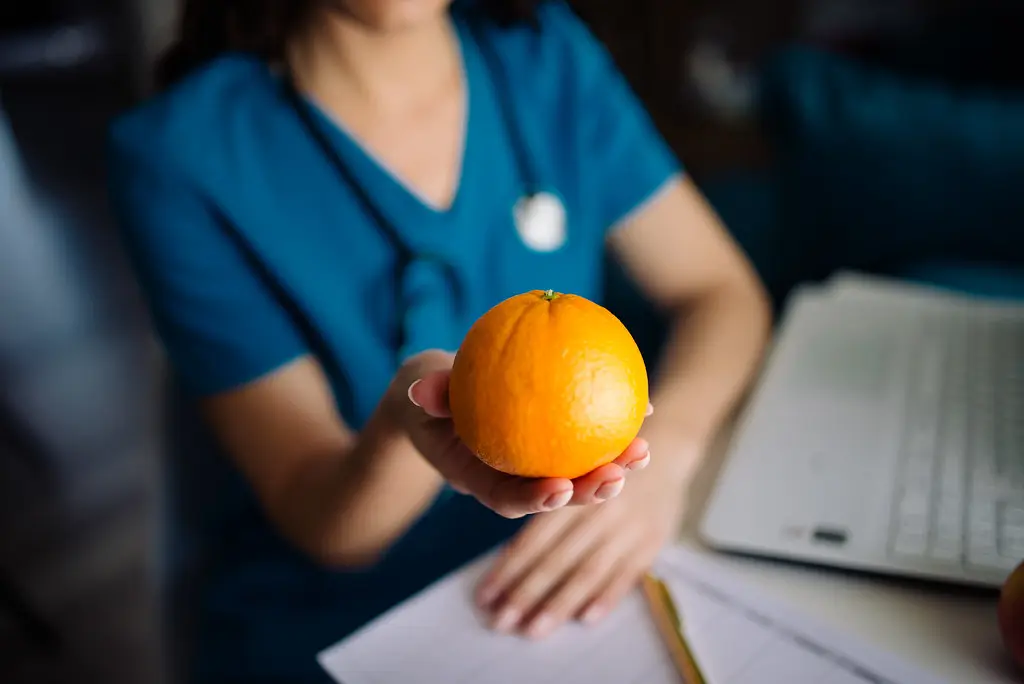 Foto: Healthcare worker presents an orange while sitting at a desk with paperwork and a laptop in a professional setting