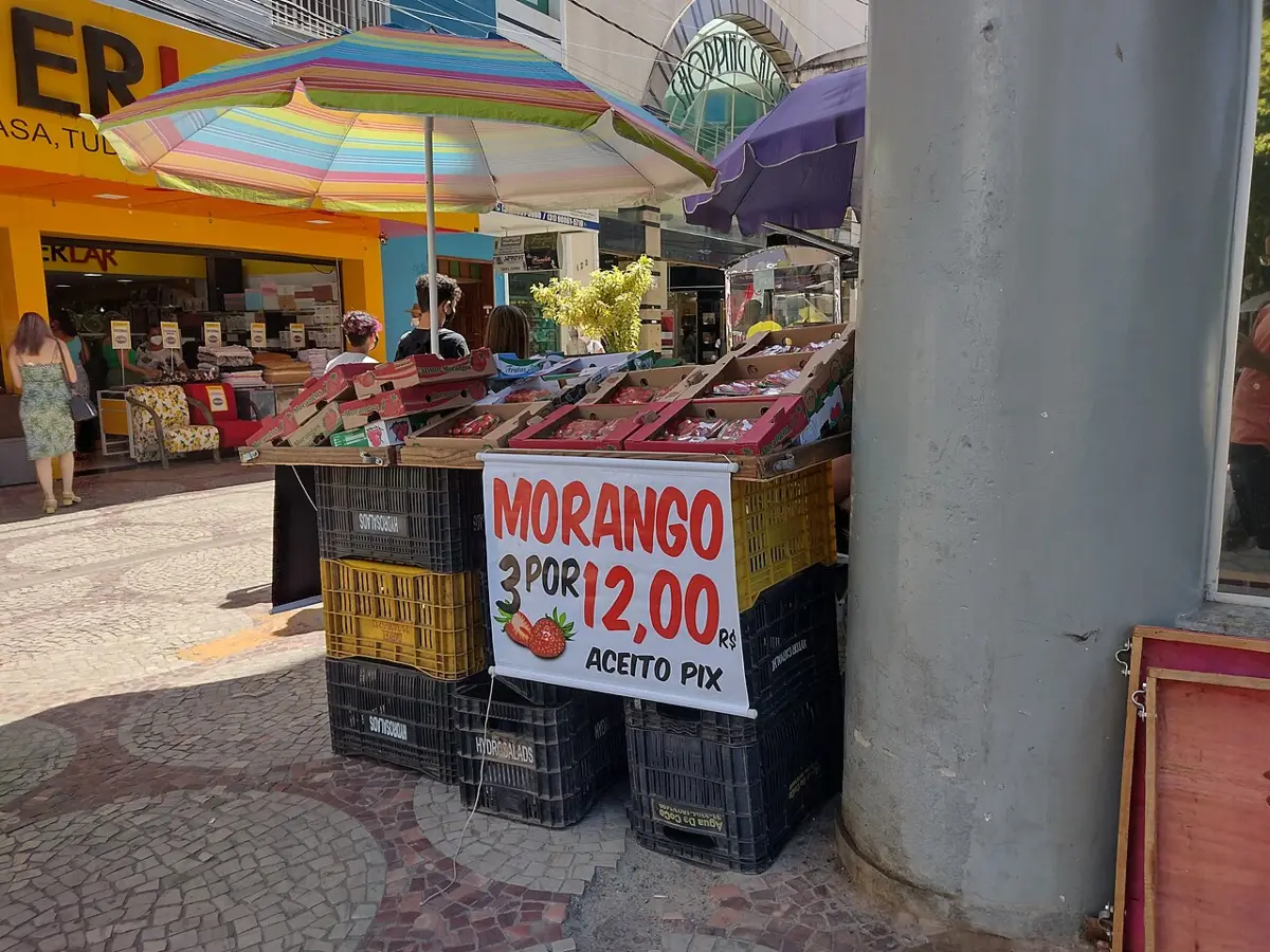 Foto: Banca improvisada feita de caixotes de plástico usada para vender morangos no calçadão central de Viçosa MG Brasil. Na placa se lê "Morango …