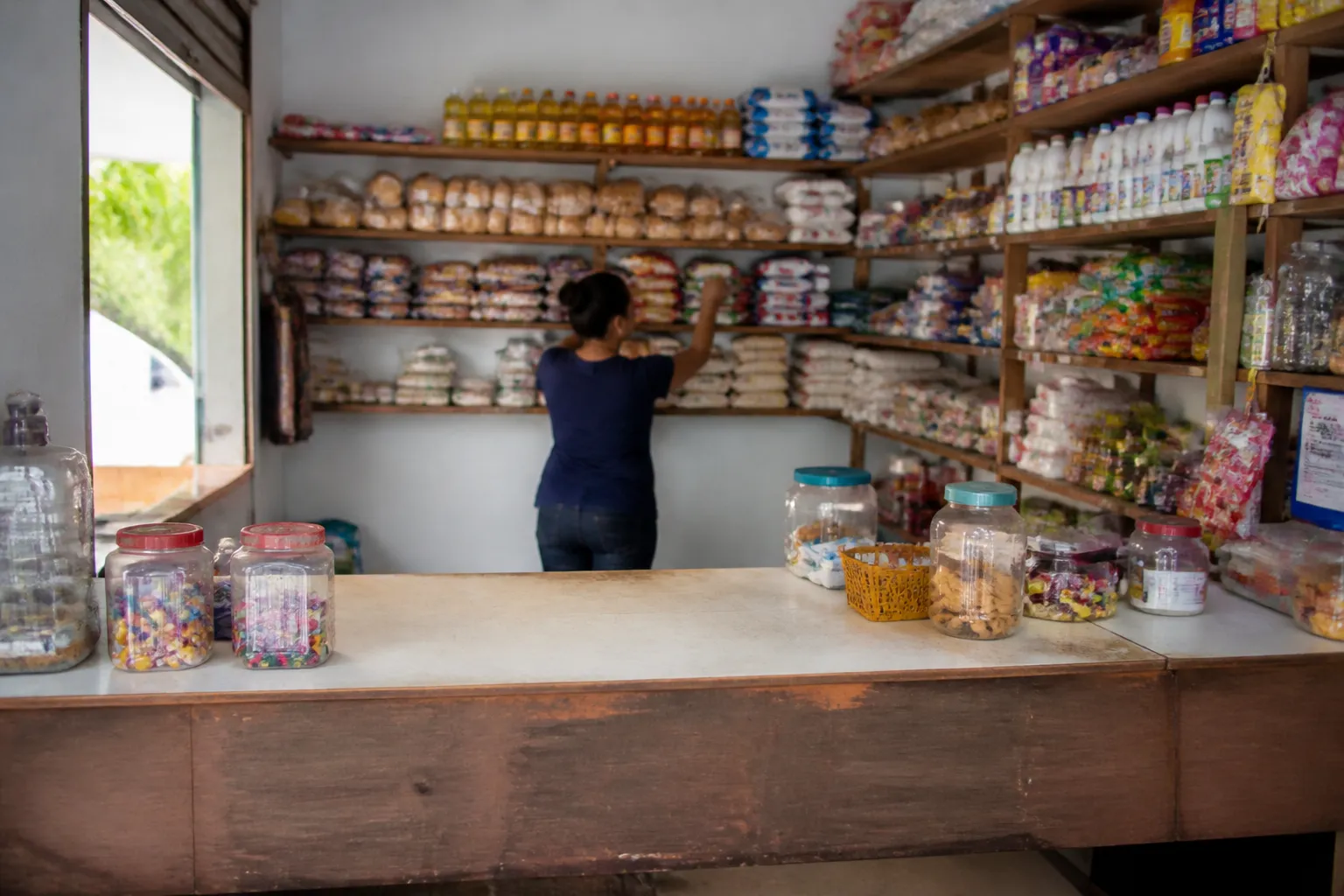 Imagem gerada por IA: Interior de uma pequena loja de bairro, com pessoa anônima organizando mercadorias.